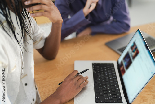 Two people are collaborating at a wooden table. An individual with dark skin and dreadlocks actively uses a laptop, holding a pen, while viewing online video content on the screen.