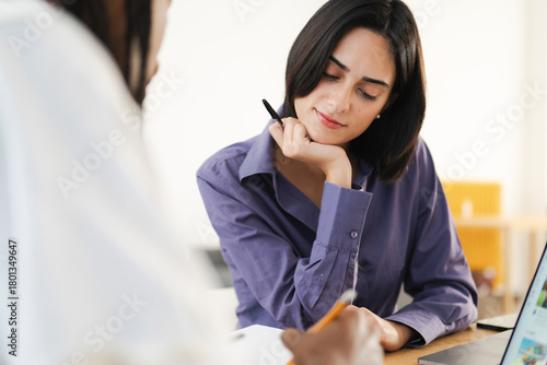 A thoughtful young woman in a purple shirt listens intently during a business meeting. Another person takes notes in the foreground. They are collaborating in an office setting.