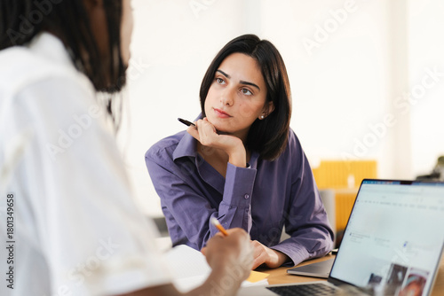 Two diverse businesswomen are collaborating in an office setting. One woman listens intently while the other takes notes during their meeting. They are focused on their work.