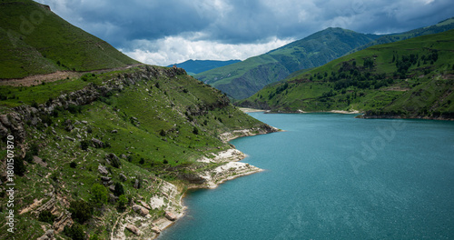 Turquoise lake nestled in green mountainous terrain, under a dramatic cloudy sky, showcasing nature's contrast between water and land