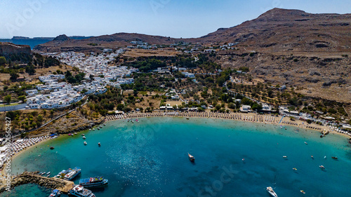 Aerial view of the sparkling turquoise bay embraces the golden sands of the beach below the whitewashed village clinging to the rugged hillside, Lindos, Rhodes, Greece.