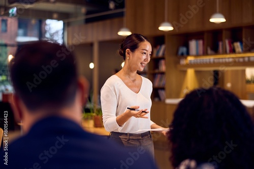 Asian Businesswoman Presenting In Modern Office Meeting With Colleagues And Bookcase Background