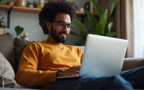 Indian man wearing glasses using laptop while sitting on couch at home. High quality