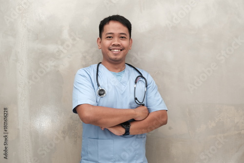 Happy Southeast Asian male nurse in scrubs with stethoscope, arms crossed, isolated on wall, ideal for healthcare ads and medical content.
