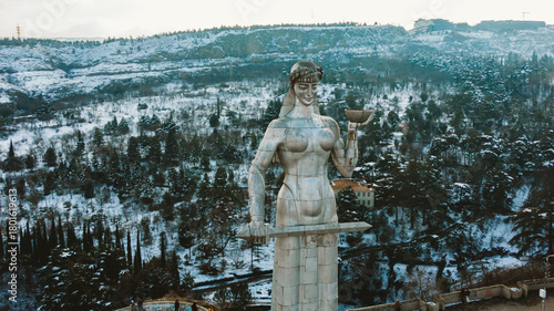 Kartlis deda statue standing high above tbilisi, embodying georgian spirit with welcoming sword and bowl, overlooking snow-covered urban panorama against mountainous backdrop