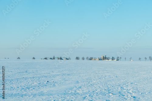 Expansive winter landscape featuring a serene snowy field under a clear blue sky, with distant buildings and trees creating a tranquil rural scene with copy space