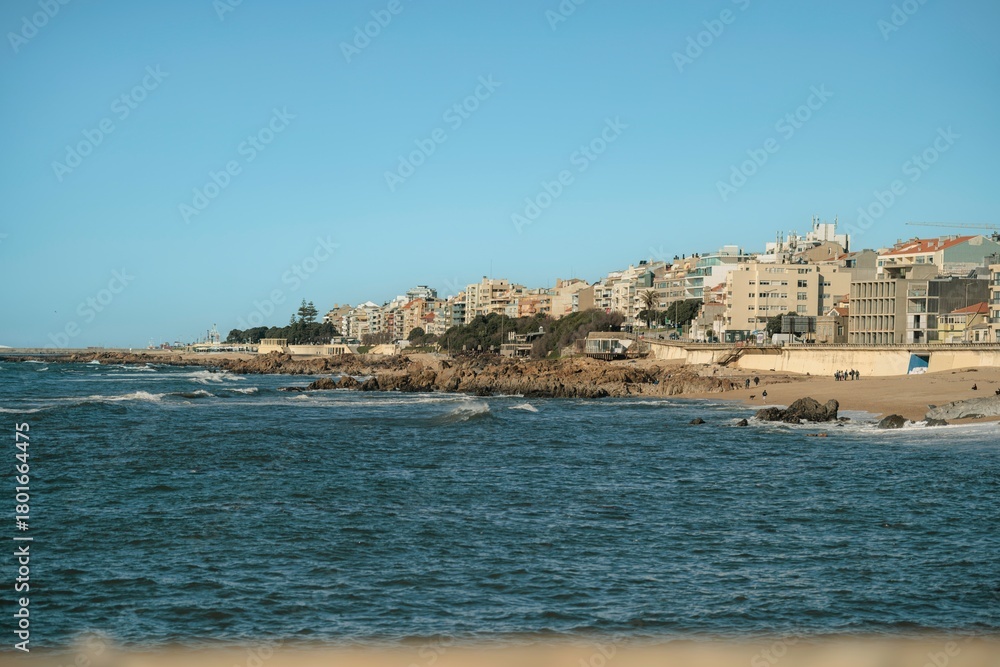 Fototapeta premium Early morning light shines on a vibrant coastal city. Waves gently crash against the rocks as buildings rise against the backdrop of a blue sky, creating a serene atmosphere