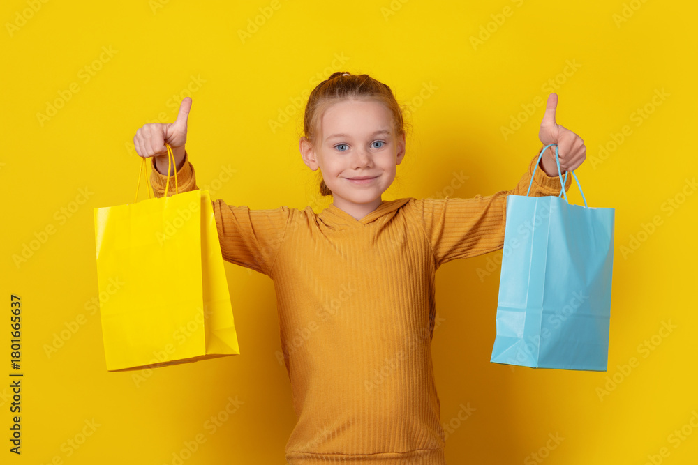 Fototapeta premium Smiling young girl in hoodie holding colorful shopping bags and showing thumbs up against bright yellow background.