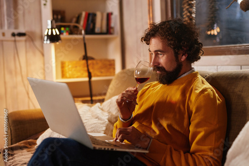 A man sits comfortably on a sofa in a warm living room, sipping red wine while focused on his laptop. The soft glow of a lamp enhances the relaxed evening atmosphere.