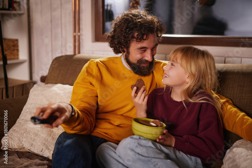 Fototapeta A smiling man and a young girl share a joyful moment while watching a movie on the couch