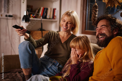 A family gathers in their warm living room for a movie night. Laughter fills the air as the parents and their child enjoy the film together, cuddled up on the couch with snacks.