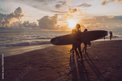 Group of women carry a longboard along the shoreline at sunset, shadows stretching across wet sand as swells roll in. Concept of teamwork, shared learning, and community powered confidence