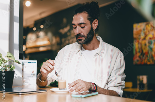 Man stirring coffee at cafe table with smartphone beside, focused moment, casual attire, relaxed remote worker enjoying balance of technology and personal lifestyle.