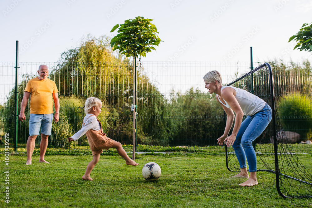 Naklejka premium Little boy playing football with mother and grandfather in backyard.