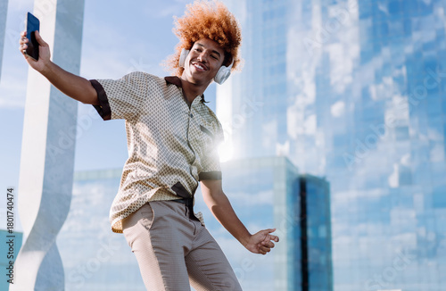 Confident African American man with red afro wearing wireless headphones, smartphone in hand, enjoying music, symbolizing personal freedom, relaxation, and technology-driven lifestyle outdoors.