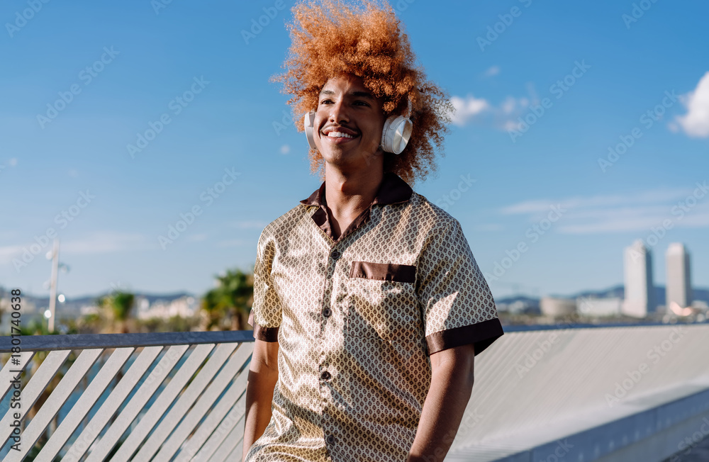 Fototapeta premium Stylish man with curly ginger afro and wireless headphones walking confidently, touching his hair, symbolizing individuality, personal technology, and the joy of music in motion.