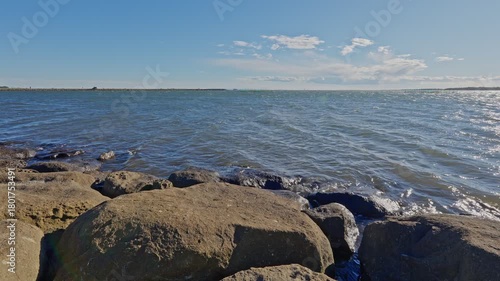 A wide shot of the vast, choppy water of Tokyo Bay, framed by large, dark rocks in the foreground under a bright sky.