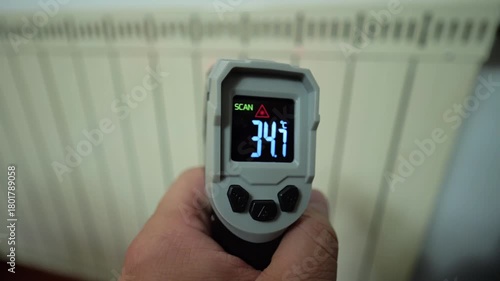 A hand holding an infrared thermometer aimed at a white radiator, demonstrating the process of checking thermal efficiency for central heating