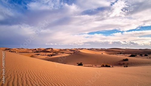 Fototapeta Naklejka Na Ścianę i Meble -  A wide, sweeping view of a desert landscape dominated by large, undulating sand dunes under a dynamic sky with scattered clouds and patches of blue.