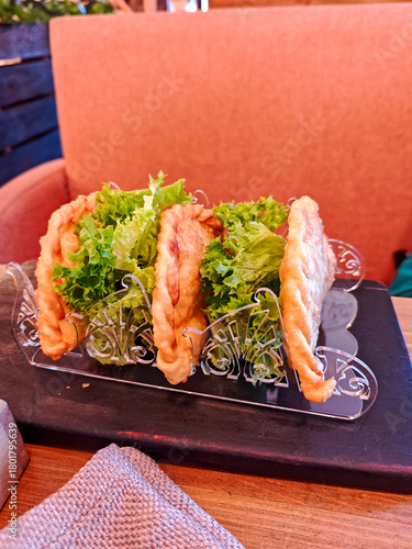 Crispy fried chebureki, traditional Eastern European meat turnovers, served with fresh lettuce in a metal holder on a wooden table in a cozy restaurant setting.