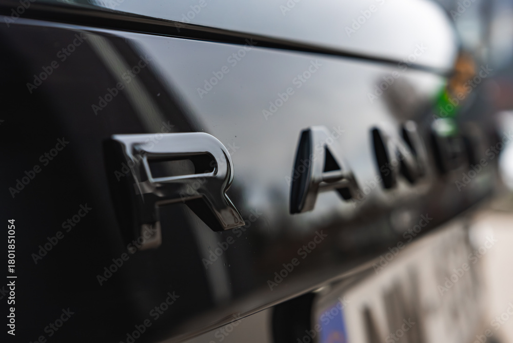 Obraz premium Close view of Range Rover rear letters in focus, glossy black paint reflects light. Soft blur hides other letters in a well lit workshop with luxury models nearby.