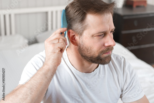 Handsome man combing brushing hair sitting at home light white bedroom. Guy styling grooming his hair with a comb. Self-care. Softly lit indoor space. Portrait. Male haircare morning routine concept