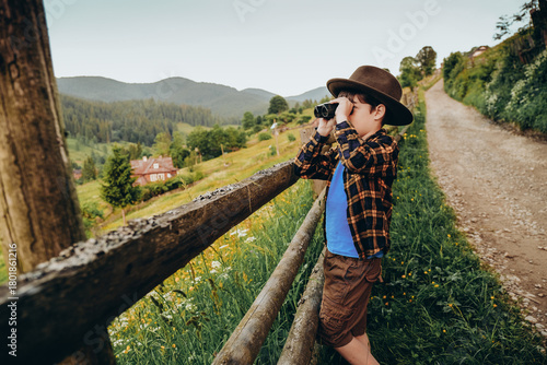 A boy looks through binoculars while standing on a hilltop in summer. Family trip to the mountains in summer.