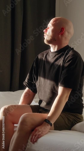 Vertical video of a man waking up in a softly lit bedroom, removing the blanket and stretching while sitting on the bed. A calm morning routine moment at home. High quality 4k footage