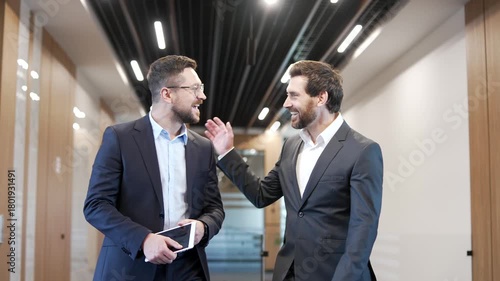 Two smiling businessmen engaging in casual discussion while walking along corridor of businss center. Happy colleagues have friendly conversation, talking. Teamwork, communication, collaboration