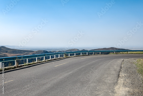 Curving asphalt highway on the edge of a mountain overlooking a valley under a clear blue sky.