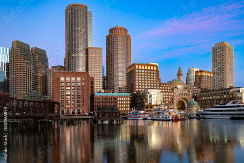 A stunning view of the Boston downtown skyline at twilight, seen across the water from the Seaport District, with brightly lit towers and colorful reflections.