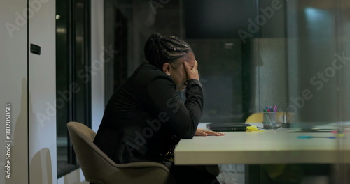African American businesswoman sits in silence at office table with hand over face, processing emotional stress and fatigue after long and intense workday