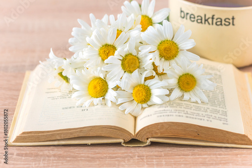 Summer still life; bouquet of white daisy or chamomile flowers on a book and cup of coffee on a wooden table; close up