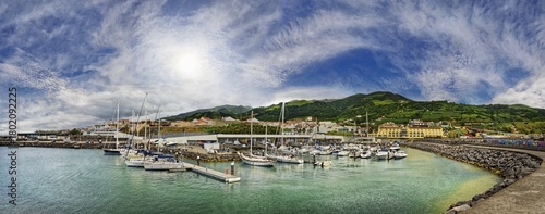 A picturesque harbour with yachts and boats, surrounded by buildings, mountains and a cloudy sky with sun, all in an impressive panoramic shot, small town of Vila Franca do Campo, Sao Miguel, Azores, Portugal
