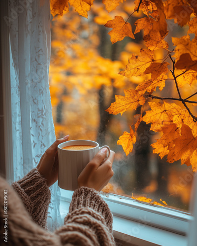 a person holding a warm drink while looking out the window at orange leaves dri