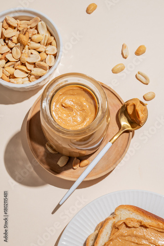 Peanut butter in a glass jar on a light background with scattered nuts, spoon and toast.