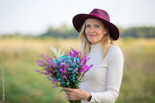 Woman in purple hat holds colorful flower bouquet in sunny field with greenery in background