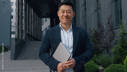 Asian middle-aged businessman with laptop smiling Korean business man posing in city holding computer device smile happy Japanese Chinese executive employer corporate company worker looking at camera