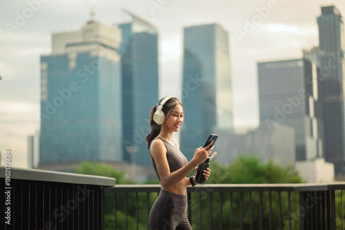 Asian woman wearing headphones standing outdoors in an urban city environment, happily checking her smartphone while holding a water bottle after a workout session.
