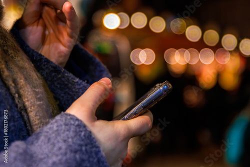 A closeup of hands holding a smartphone against a blurred background of warm festive lights at a night holiday market. Selective focus
