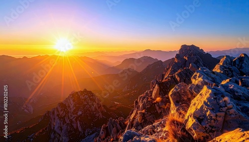 A vibrant sunrise casts golden light across layers of rugged mountain peaks under a clear blue sky.