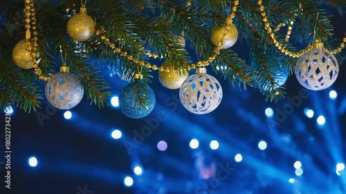 A festive close-up video of golden Christmas ornaments hanging on a fir tree branch, set against a vibrant blue bokeh background. The sparkling decorations and soft glowing lights create a warm and el