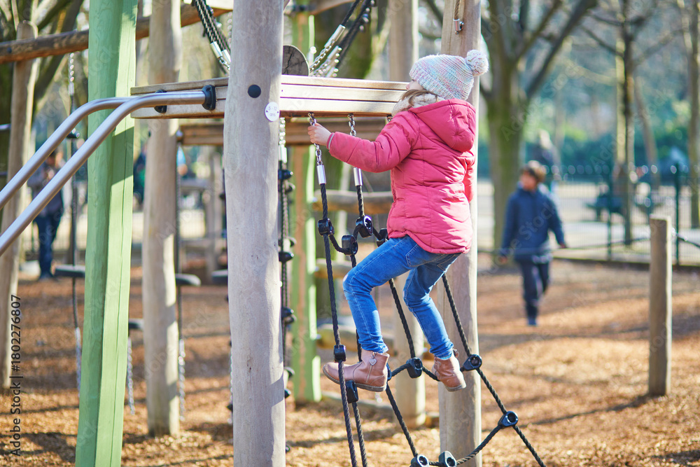 Fototapeta premium A young girl in warm winter clothing balancing and climbing on wooden playground equipment outdoors
