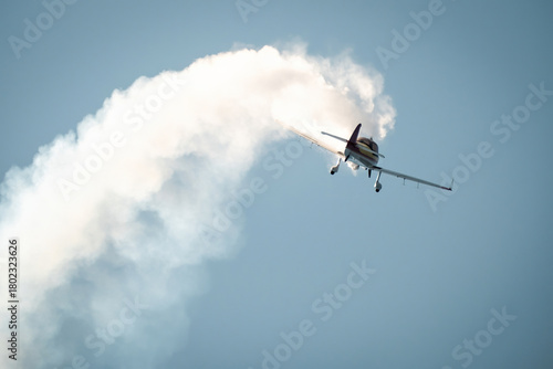 Small aerobatic aircraft flying upward leaving a thick white smoke trail during an air show against a bright blue sky.