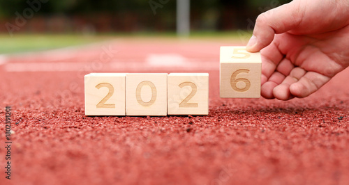 A hand places a wooden block showing the number six next to blocks showing 2026, symbolizing the transition to the new year. A joyful atmosphere celebrates the upcoming year