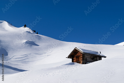 Wooden alpine cabin in snowy mountain landscape with ski tracks in the background under deep blue sky