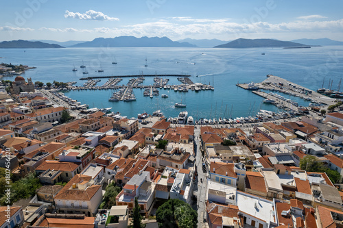 Fototapeta Naklejka Na Ścianę i Meble -  Aerial view of Aegina town and its marina in Greece, showcasing terracotta rooftops, sailboats, and the surrounding Saronic Gulf islands under clear Mediterranean skies.