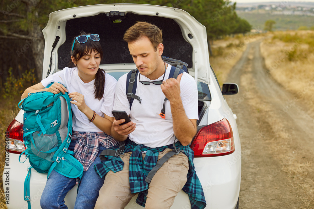 Obraz premium Couple by car use phone, map, backpack. Stranded on a rural road, travelers check geolocation, seek signal, and study a map while resting on the trunk near the forest. Concept: lost on the road.
