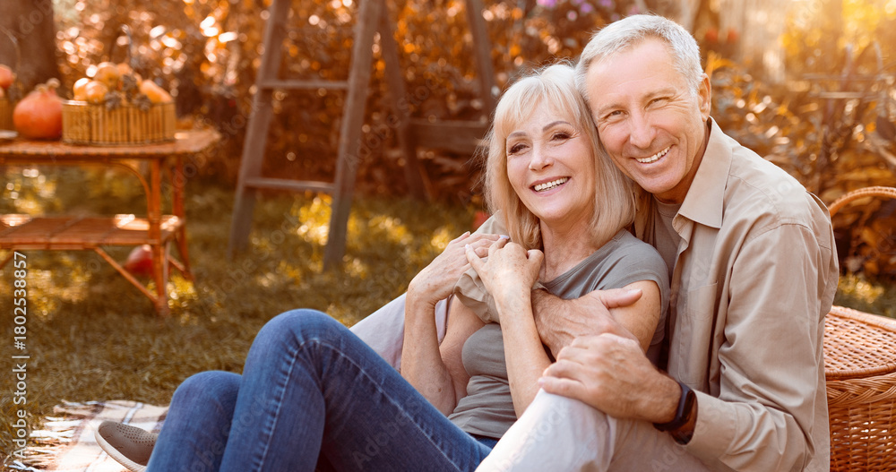 Fototapeta premium A joyful couple sits together in their backyard, surrounded by autumn leaves and pumpkins. They share a warm embrace and smile at each other, enjoying the cozy atmosphere of the season.