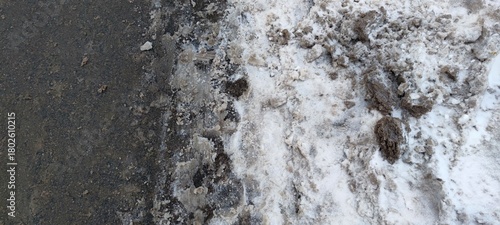 The edge of road. A winter day. The asphalt road is covered in dirty, black-brown snow. A small drift of white snow lies at the edge. Clumps of dirty, sticky snow are visible throughout the snowdrift.
